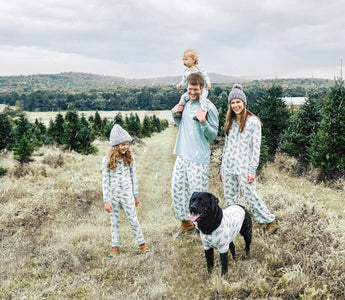 A family of 4 with a dog standing outside in matching pajamas at a Christmas tree farm.